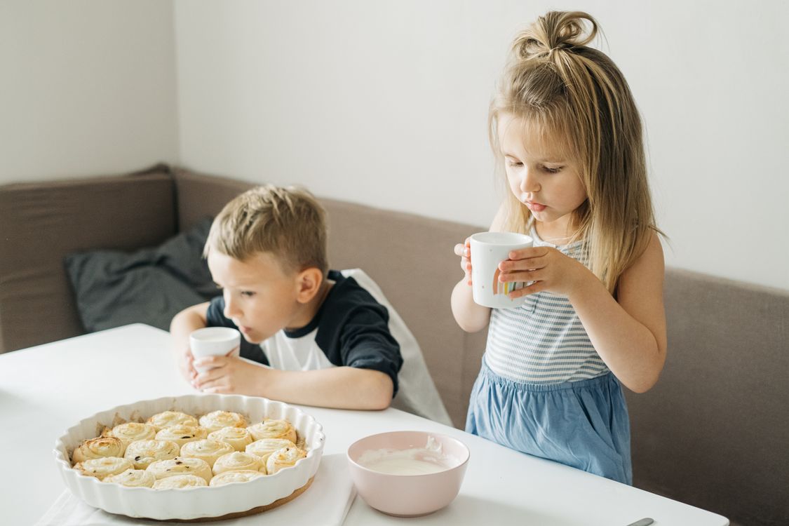 Två barn som dricker mjölk till kanelbullar vid köksbordet